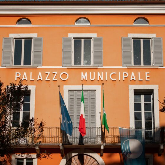 Orange town hall facade with three arched windows and gray shutters, flags hanging below.