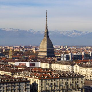 Turin skyline with the Mole Antonelliana tower rising above red rooftops and snow-capped Alps on the horizon.