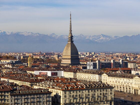 Turin skyline with the Mole Antonelliana tower rising above red rooftops and snow-capped Alps on the horizon.