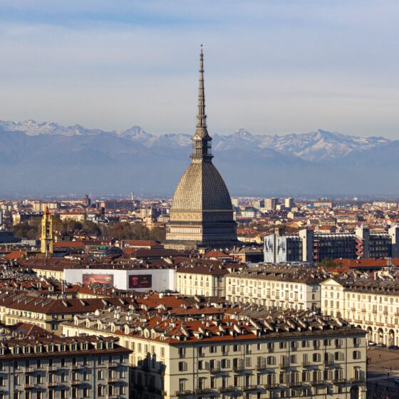 Turin skyline with the Mole Antonelliana tower rising above red rooftops and snow-capped Alps on the horizon.