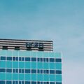 Corner of a glass hotel with turquoise windows and a blue 'HOTEL' sign against a clear sky.