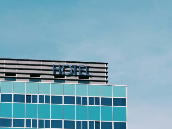 Corner of a glass hotel with turquoise windows and a blue 'HOTEL' sign against a clear sky.