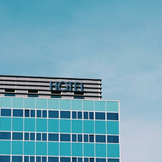 Corner of a glass hotel with turquoise windows and a blue 'HOTEL' sign against a clear sky.